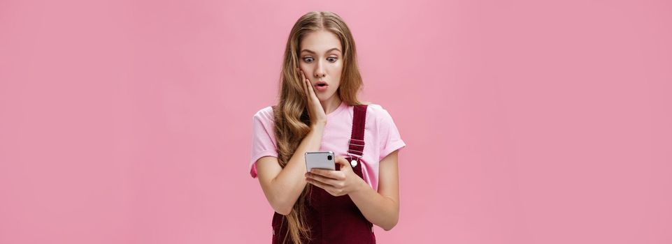 Girl found out shocking truth after reading message in smartphone pressing arm to cheek in surprise staring concerned and speechless at cellphone screen reacting to unexpected news over pink wall
