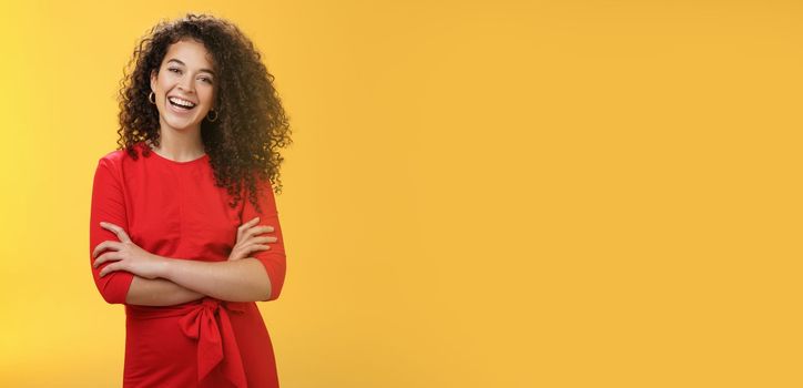 Self-assured happy enthusiastic curly-haired female reporter in cute red dress laughing carefree, having fun tilting head amused and holding hands crossed over body in confident pose over yellow wall