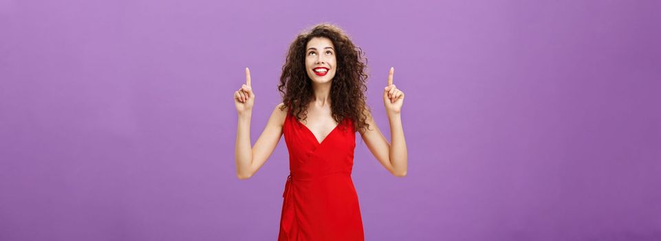 Woman going on street breat fresh air and enjoy fireworks in her honor looking and pointing up with dreamy, delighted expression smiling joyfully standing over purple background in red evening dress