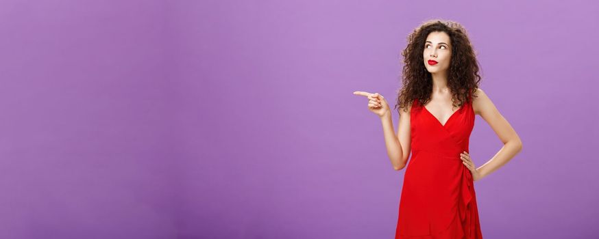 Portrait of thoughtful and dreamy charming woman with curly hairstyle in elegant red evening dress pointing and looking left curiously holding hand on waist posing feminine over purple wall