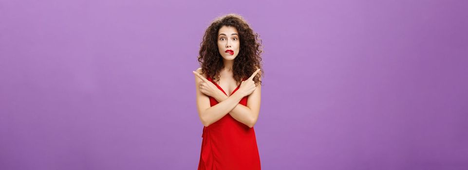 Enthusiastic unsure emotive elegant woman in stylish evening red dress biting lip looking excited pointing in different directions left and right being uncertain and doubtful in making decision