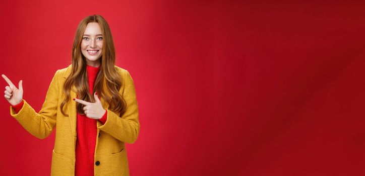 Cheerful friendly-looking and energized helpful redhead girl in yellow coat showing way pointing at upper left corner and smiling broadly with satisfied grin posing delighted over red background