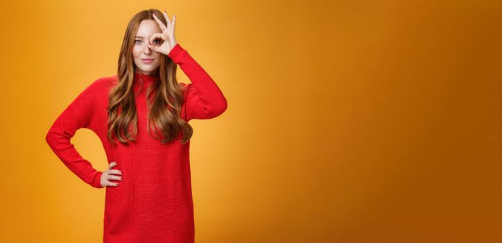 I got you on aim. Self-assured expressive and confident attractive redhead woman in red warm dress showing okay or zero sign on eye and peeking through as smiling satisfied over orange wall