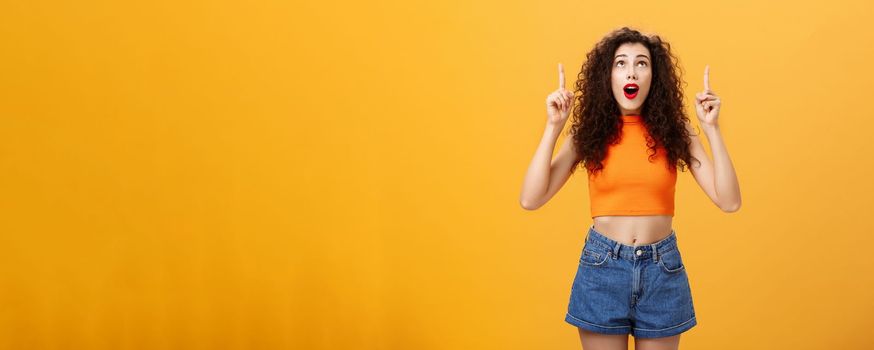 Portrait of amazed emotive and charismatic young curly-haired woman with red lipstic looking and pointing up mesmerized and astonished standing under impression over orange background