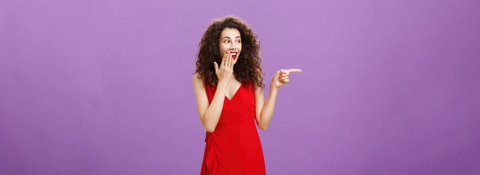 Girl chuckles over clumsy guy making mistake during party covering smile with palm gazing and pointing left while gossiping with girlfriends joyfully standing in elegant red dress over purple wall