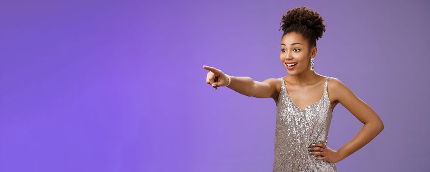 Impressed fascinated cute african-american woman excited pointing index finger sideways thrilled enjoying incredible exhibition standing excited blue background, glancing amused