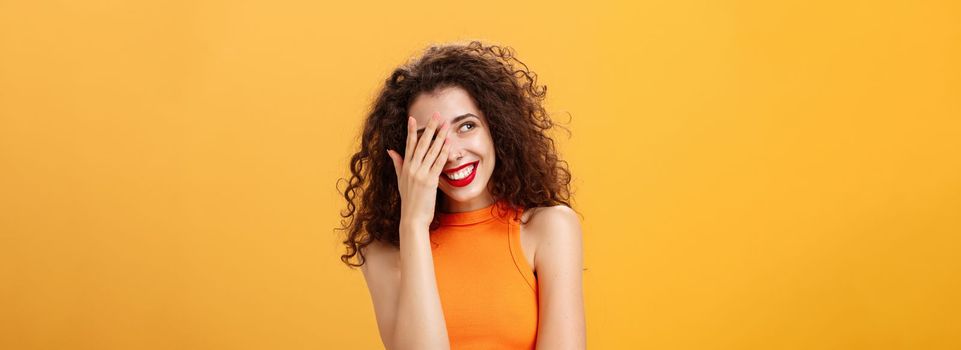 Woman being praised by coworkers feeling awkward and shy hearing congratulations with received award covering face peeking left with joyful satisfied smile posing over orange background