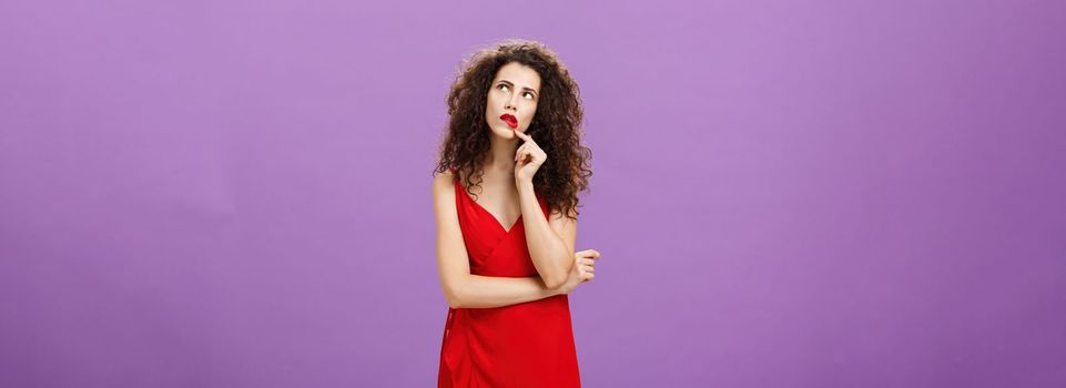 Elegant thoughtful woman in trendy red dress with curly hairstyle and party makeup touching lip looking at upper right corner thinking how make impression during conversation over purple background