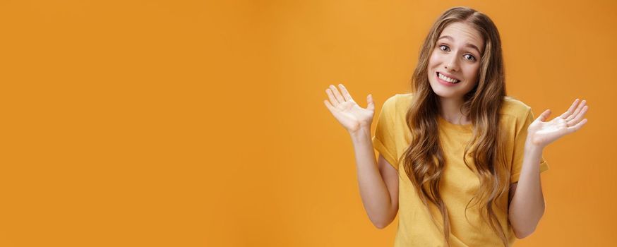 Girl stay out of business shrugging with hands raised and spread aside, silly sorry smile posing over orange background unaware and confused in casual t-shirt having no idea about topic