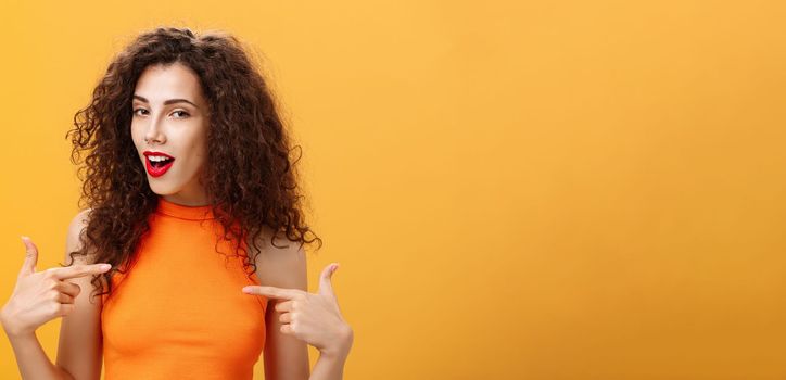 Proud and satisfied cool urban female with red lipstick and curly hairstyle pointing at herself with self-assured expression winking bragging about skills and achievements over orange background