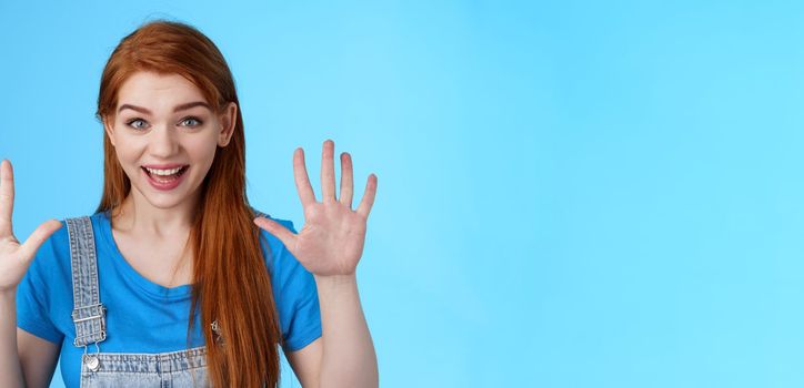 Close-up friendly joyful redhead caucasian girl, long ginger hairstyle, raise hands show number ten, counting dozer, explain fingers, raise arms surrender, smiling joyful, stand blue background