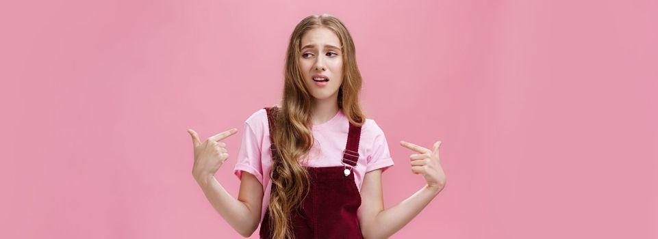 Self-assured arrogant young female student with high ego standing cocky and brag about herself pointing at her with cool snobbish look looking away to right with contempt posing over pink wall