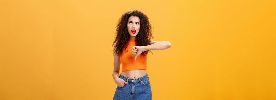 Party for losers I am out. Dissatisfied attractive popular girl with curly hairstyle in red lipstick and cropped top rolling eyes up from irritation and dislike showing thumb down over orange wall