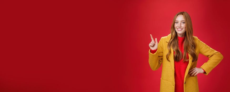 Charming young female in yellow coat holding hand on waist and pointing at upper left corner, smiling broadly as showing or promoting interesting proposal over red background, in good mood