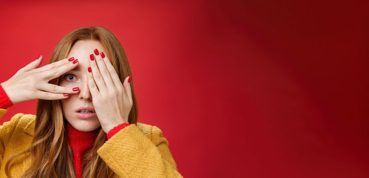 Stunned and shocked redhead woman cannot believe what terrible thing she sees covering face with palms open mouth from shook and peeking through fingers anxious and scared over red background