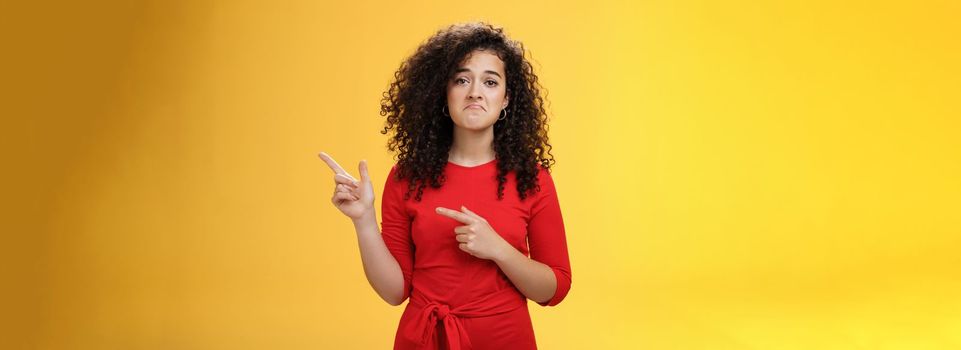 Waist-up shot of upset displeased girl with curly hair being down in dumps pointing at upper right corner with disappointed gloomy smile, feeling regret, sadness standing like loser over yellow wall