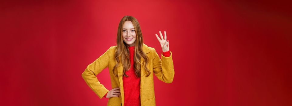 Cute friendly-looking ginger girl showing three reasons buy product smiling broadly making third reservation, looking delighted and confident in her choice posing against red background in yellow coat
