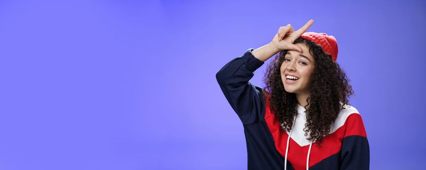 Girl triumphing and mocking friend as winning showing loser gesture on forehead and laughing having fun standing pleased and happy over blue background, smiling at camera joyfully