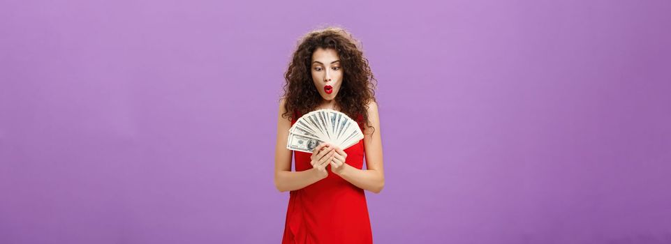 Portrait of surprised and amazed speechless woman folding lips making wow sound while holding and looking at lots of money being rich and successful receiving lots of cash over purple background