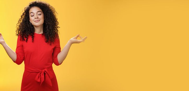 Charming dreamy and happy peaceful woman in red dress with curly hair standing in lotus pose with mudra orbs or zen gesture and closed eyes, meditation relieving stress, feeling unbothered. Body language, buddhism and people concept