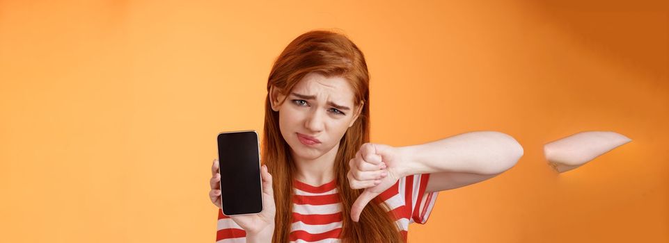 Very bad poor quality. Upset redhead female customer grimace disappointed, show smartphone blank display, thumbs down judgement, dislike app, smirk dismay, stand upset orange background