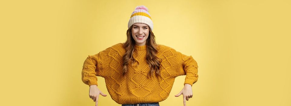 Friendly-looking helpful good-looking 20s college student girl wearing hat warm sweater smiling pleased showing you awesome promotion pointing index fingers down recommend bottom advertisement