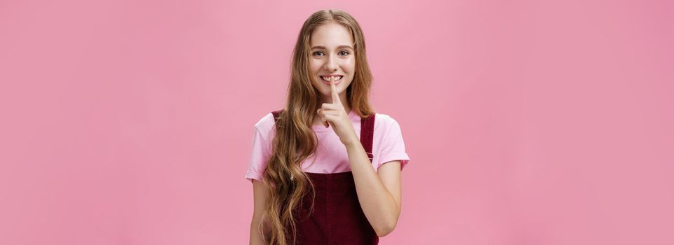 Girl wanna share her beauty secret smiling tricky and friendly showing shush gesture with index finger over mouth to save surprise and mystery posing delighted and relaxed over pink background