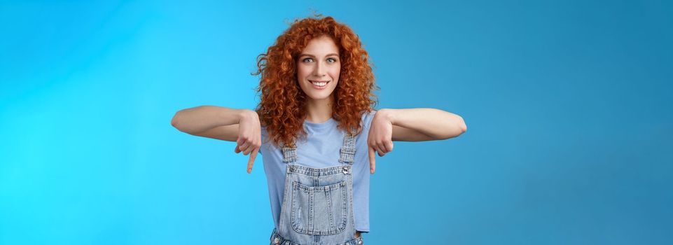 Charismatic sassy flirty redhead daring ginger girl curly haircut pointing down index fingers smiling broadly enthusiastic explore new store pointing promo like cool advertisement blue background