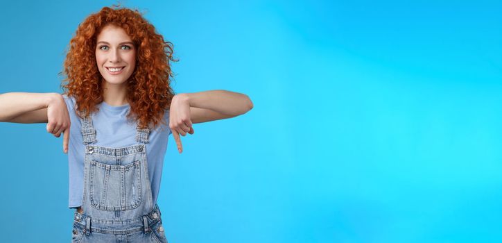 Charismatic sassy flirty redhead daring ginger girl curly haircut pointing down index fingers smiling broadly enthusiastic explore new store pointing promo like cool advertisement blue background