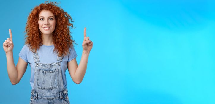 Motivated happy cheerful redhead silly curly woman pointing up inrdex finger smiling charmed impressed excited showing awesome promo discuss interesting advertisement blue background