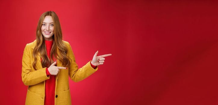 Carefree cute and tender young redhead female student in stylish yellow coat over dress pointing right and smiling with delighted and impressed look showing great place over red background