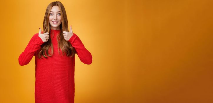 Excited and supportive redhead girlfriend asking opinion about new warm dress showing thumbs up and raising eyebrows questioned as waiting for opinion over orange background