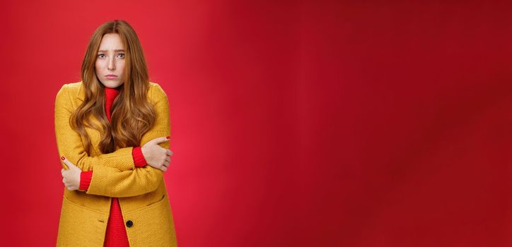 Cute and sad trembling redhead girl in light yellow coat shaking from cold, having goosebumps frowning and pouting from sorrow as freezing on windy weather over red background. Copy space