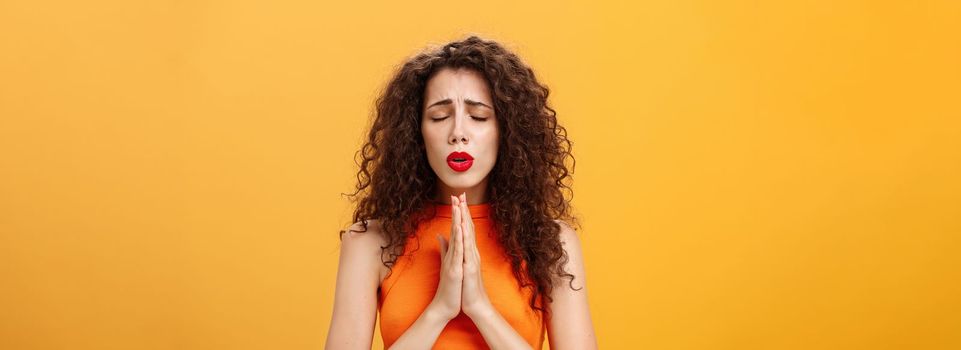 Nervous and concerned woman with curly hairstyle feeling hopeful praying with closed eyes and frowned eyebrows holding hands in pray near chest hopefully dreaming troubled will solve over orange wall