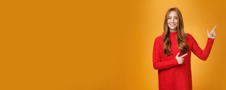 Friendly-looking joyfuly and energized redhead female in red sweater pointing at upper right corner promoting advertisement with broad delighted and tender smile posing against orange background