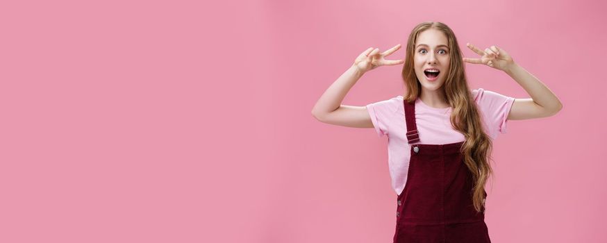 Girl staying positive lifting after falling. Charming carefree friendly-looking young woman with small scar on arm and tattoo showing peace gestures, open mouth and looking at camera over pink wall