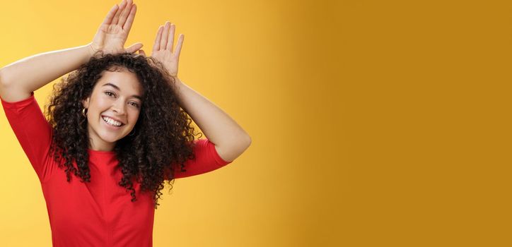 Close-up shot of charismatic playful and tender young kind woman with curly hair playing having fun showing bunny ears with hands on head tilting head joyfully fooling around and smiling at camera