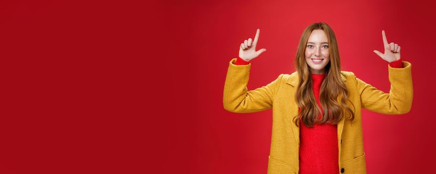 Indoor shot of redhead attractive woman in yellow fall coat raising hands promoting advertisement as pointing up and smiling broadly with satisfied pleasant expression over red background