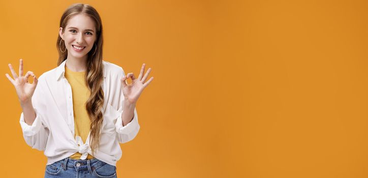 Indoor shot of assertive and assured charming stylish woman in blouse over t-shirt and accessories showing okay gesture with delighted self-assured smile giving being ok against orange wall