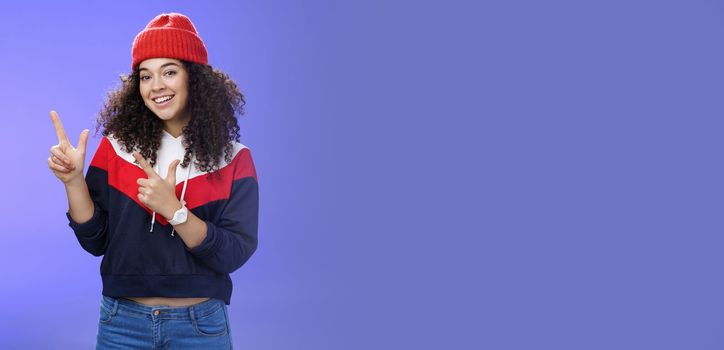 Portrait of friendly good-looking woman with curly hair in red warm beanie pointing at upper left corner and smiling delighted as promoting cool copy space against blue background