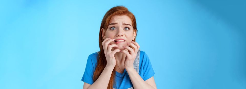 Close-up intense panicking redhead woman watching scary movie feel pressured, biting lip worried, hold hands near mouth self-soothing trying calm down, stare camera anxious, blue background