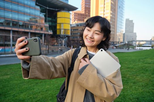 Happy university student, korean girl takes selfie with her papers and digital tablet, holds smartphone and poses near university campus