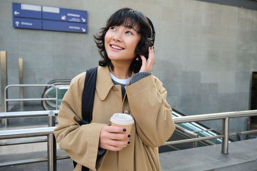 Portrait of young student, girl in headphones, drinks coffee, stands on street with backpack, commutes to university or college, smiles happily