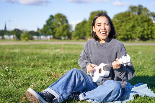 Portrait of beautiful asian woman singing, playing ukulele guitar in park, sitting alone on grass on sunny day