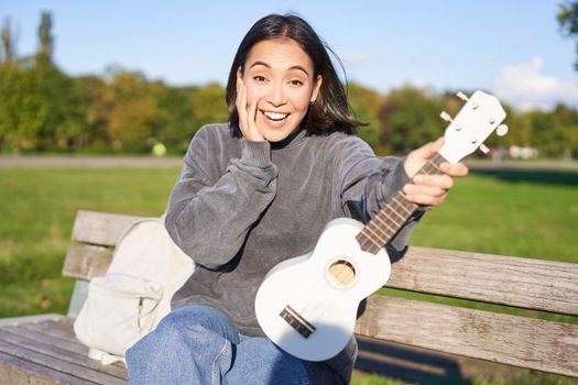Portrait of young girl musician, sitting in park with ukulele guitar, looking surprised at camera, saying wow
