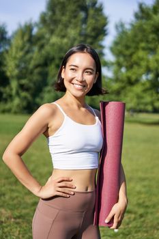 Vertical portrait of young asian fitness girl walks with rubber mat for yoga, goes on training session on fresh air in park, wears sports clothing