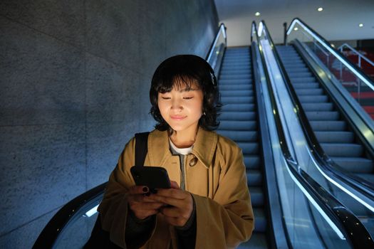 Cute young woman going down the escalator to the tube, using subway metro to commute to work or university, standing with smartphone