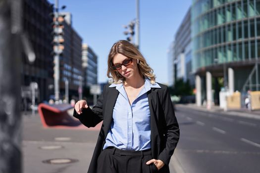 Portrait of confident corporate woman, young saleswoman in suit and sunglasses, looking self-assured on city street
