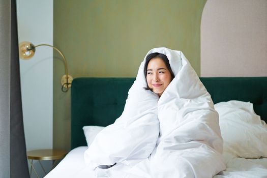 Cozy mornings. Happy asian girl feels warm in her bed, covers herself with cozy duvet in her bedroom, a comfortable stay in hotel room