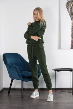 Stylish beautiful young blond woman in a green tracksuit poses near a white wall in the room. Attractive girl model posing near blue chair.